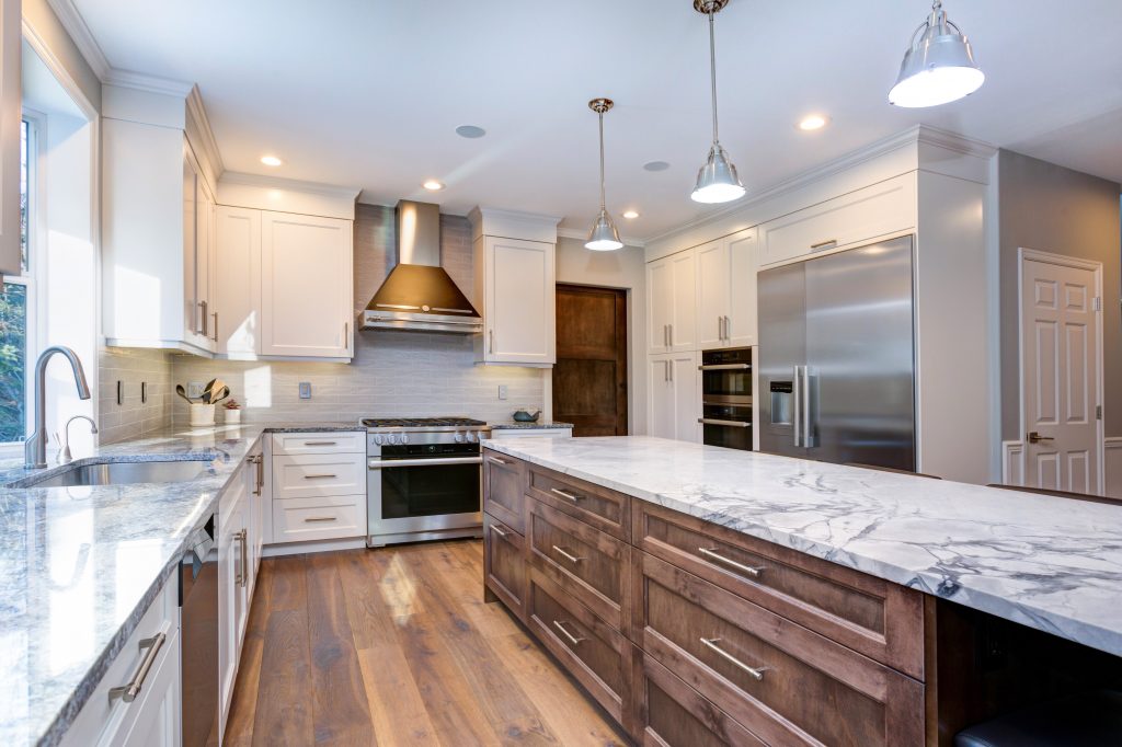 Luxury home interior boasts amazing white kitchen with custom white shaker cabinets, endless marble topped kitchen island with drawers and stainless steel appliances over wide planked hardwood floor.